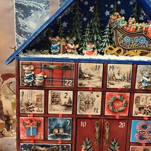 Puede incluir: Un calendario de adviento de madera con forma de casa y techo azul, que cuenta con veinticuatro cajones numerados decorados con ilustraciones festivas de ratones, Pap&aacute; Noel, paisajes invernales y vegetaci&oacute;n, coronado con una escena de trineo.