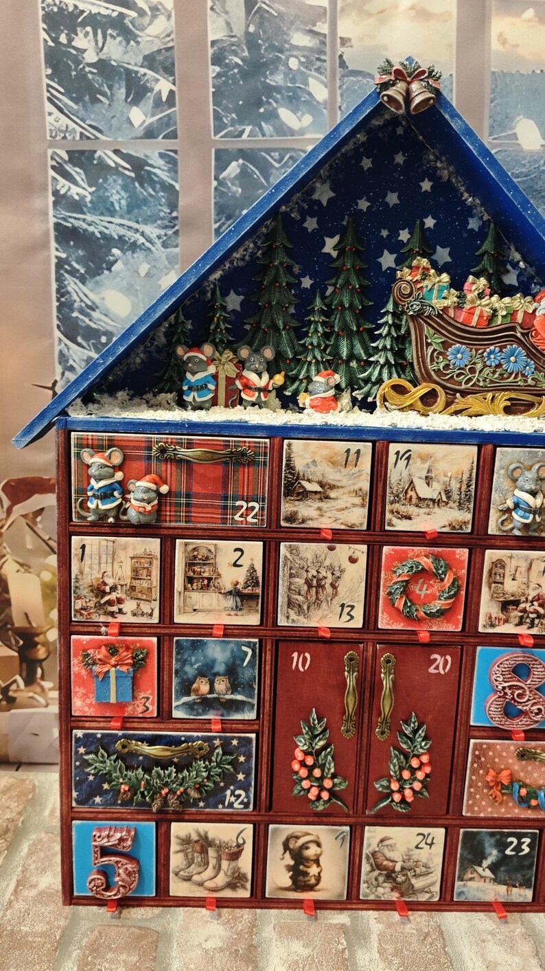 Puede incluir: Un calendario de adviento de madera con forma de casa y techo azul, que cuenta con veinticuatro cajones numerados decorados con ilustraciones festivas de ratones, Pap&aacute; Noel, paisajes invernales y vegetaci&oacute;n, coronado con una escena de trineo.