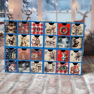 Puede incluir: Un calendario de Adviento de madera rectangular con veinticuatro cajones pequeños, cada uno con una ilustración navideña única y un número. El marco está pintado de azul y los frentes de los cajones muestran imágenes festivas que incluyen ratones, Santa Claus y coronas.