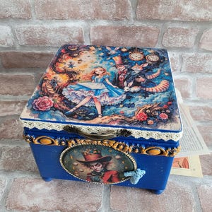 Puede incluir: Una caja de madera azul con temática de Alicia en el país de las maravillas. La tapa presenta una ilustración colorida de Alicia, el Sombrerero Loco y el Gato de Cheshire. La caja tiene un borde de encaje y un retrato del Sombrerero Loco.