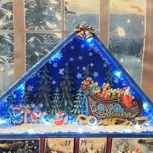 Puede incluir: Un calendario de adviento de madera triangular con techo azul que presenta una escena invernal en miniatura con un trineo de Santa, &aacute;rboles de hoja perenne y figuras de ratones. La pantalla est&aacute; iluminada por luces LED y cajones numerados.