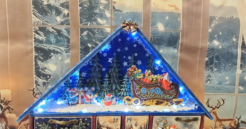 Puede incluir: Un calendario de adviento de madera triangular con techo azul que presenta una escena invernal en miniatura con un trineo de Santa, &aacute;rboles de hoja perenne y figuras de ratones. La pantalla est&aacute; iluminada por luces LED y cajones numerados.