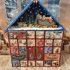 Puede incluir: Un calendario de adviento de madera con forma de casa y techo azul, que cuenta con veinticuatro cajones numerados decorados con ilustraciones festivas de ratones, escenas invernales y motivos navide&ntilde;os, rematado con un trineo en miniatura.