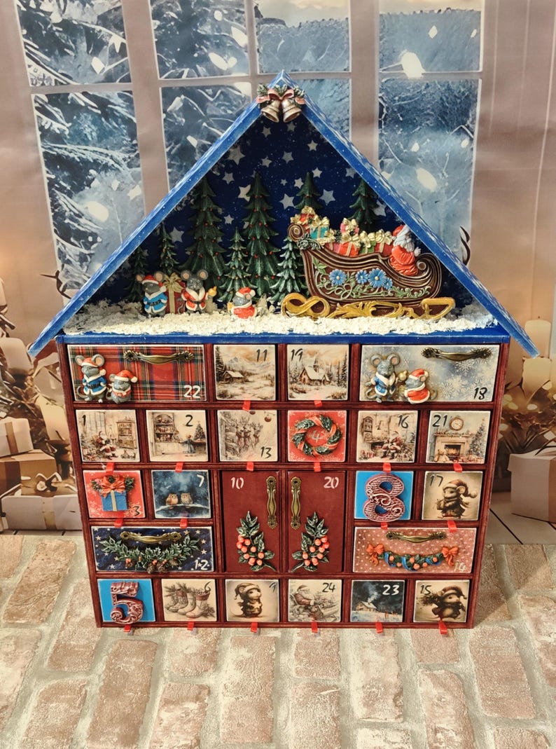 Puede incluir: Un calendario de adviento de madera con forma de casa y techo azul, que cuenta con veinticuatro cajones numerados decorados con ilustraciones festivas de ratones, escenas invernales y motivos navide&ntilde;os, rematado con un trineo en miniatura.