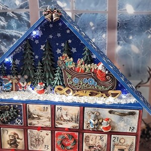 Puede incluir: Un calendario de adviento de madera con forma de casa y techo azul, que presenta una escena invernal en miniatura con un trineo, abetos y figuras de ratones. El frente tiene veinticuatro cajones numerados con ilustraciones festivas.