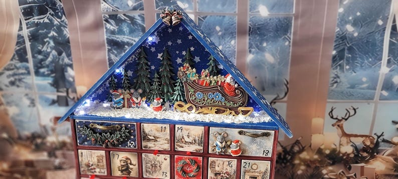 Puede incluir: Un calendario de adviento de madera con forma de casa y techo azul, que presenta una escena invernal en miniatura con un trineo, abetos y figuras de ratones. El frente tiene veinticuatro cajones numerados con ilustraciones festivas.