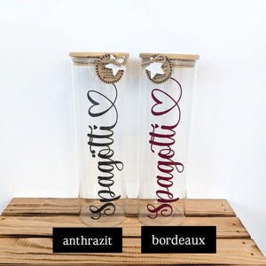 Könnte beinhalten: Zwei hohe, zylindrische Vorratsgläser aus klarem Glas mit Bambusdeckeln. Jedes Glas ist mit einem weißen Sternanhänger aus Holz an einer Schnur versehen und trägt den Schriftzug "Spagötti" in Schreibschrift mit einem Herzsymbol.
