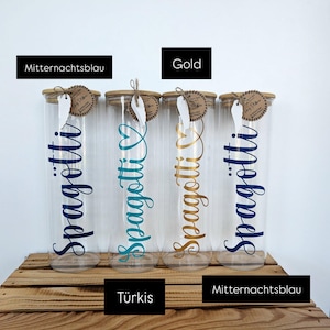 Könnte beinhalten: Vier klare Spaghetti-Gläser mit Holzdeckeln, jeweils mit "Spagotti" in verschiedenen Farben beschriftet: Marineblau, Türkis, Gold und Marineblau. Die Gläser stehen auf einer Holzoberfläche.