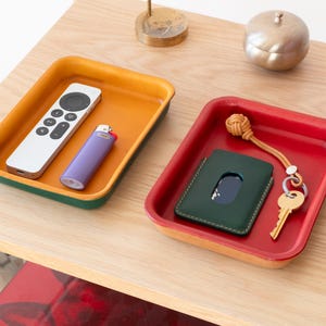 Puede incluir: Dos bandejas rectangulares pequeñas con bordes de diferentes colores. La bandeja superior es amarilla, verde y roja. Contiene un mando a distancia y un encendedor. La bandeja inferior es roja, amarilla y verde. Contiene una cartera de cuero verde, una llave con un llavero y una cuerda anudada.