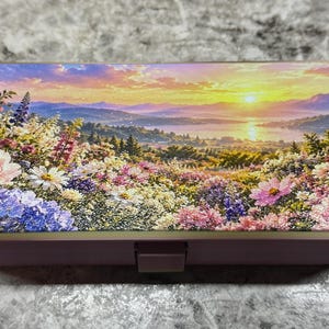 May include: A rectangular metal case with a hinged lid featuring a vibrant landscape print of a sunset over a lake and mountains, surrounded by a foreground of colorful wildflowers including pink peonies, purple lupines, and white daisies.