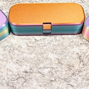 May include: Three rectangular storage cases with rounded corners and hinged lids. The two outer cases feature a textured lavender finish with a gradient transition to blue and yellow. The center case has a textured orange finish with a blue base.