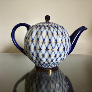 May include: A porcelain teapot featuring a white body with a blue lattice pattern and gold accents at each intersection. The teapot has a solid blue handle, a blue spout, and a matching lid with a small blue and gold knob on top.
