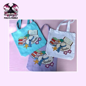 May include: Three tote bags in light blue, white, and light purple, each featuring a graphic print of a stack of books, an open book, a sun hat with a blue ribbon, a yellow starfish, a green seashell, and pink heart-shaped glasses with the word Reader.
