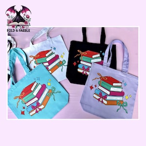 May include: Four tote bags in white, black, light blue, and lavender, each featuring a graphic illustration of a stack of four books tied with a ribbon and surrounded by small star shapes. The books are red, green, pink, and orange.
