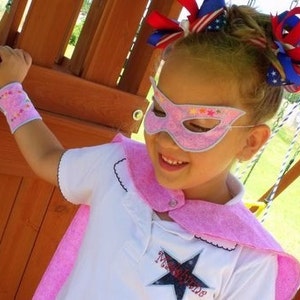 Könnte beinhalten: Ein junges Mädchen trägt einen rosa Superhelden-Umhang und eine Maske mit einem Sternmuster. Sie trägt ein weißes Shirt mit dem gestickten Text "Miss Attitude". Ihre Haare sind mit roten, weißen und blauen Schleifen gestylt.