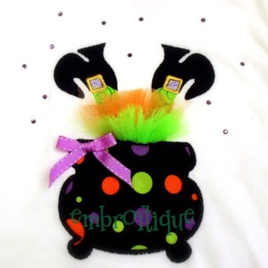 Puede incluir: Camiseta blanca con temática de Halloween con un caldero negro con lunares de colores y un lazo morado. Encima del caldero hay piernas de bruja negras con hebillas doradas y tul naranja y verde. La palabra "embrotique" está impresa en el caldero.