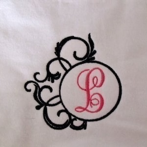 May include: A white fabric with a black embroidered design featuring a pink letter L in a circle.