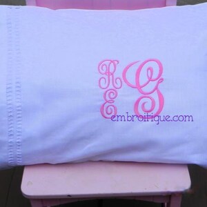 以下が含まれることがあります： ピンクの刺繍で「R.G.」のイニシャルと「embroitique.com」のウェブサイトが施された白い枕カバーです。
