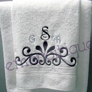 May include: White bath towel with a black and gray embroidered design. The design features a decorative flourish with a central letter "S" and two smaller letters "G" and "A" above it.