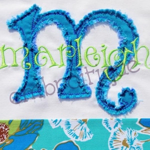 May include: A blue and green embroidered letter "M" with the name "Marleigh" stitched in green thread on a white fabric background.