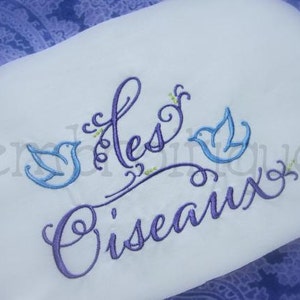 May include: Purple embroidery on a white fabric background with the text "Les Oiseaux" and two blue birds.