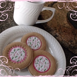 May include: Three brown felt cookies with pink and green sprinkles on a white plate. The cookies are on a white plate with a white mug in the background.