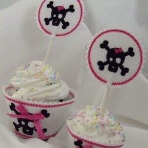 May include: Two cupcakes decorated with white frosting and colorful sprinkles. Each cupcake has a white cupcake liner with a black and pink skull and crossbones design. The cupcakes are topped with pink and white skull and crossbones toppers.