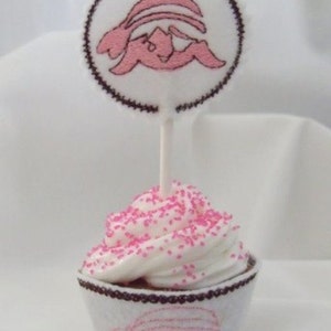 May include: A pink cupcake with pink sprinkles topped with a white cupcake topper featuring a pink embroidered bunny rabbit design.