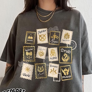 May include: Dark grey t-shirt with a gold-colored stamp design featuring fantasy class names like "Wizard," "Fighter," and "Druid." The shirt has a relaxed fit and the text "COMFORT COLORS Pepper" in a white oval.