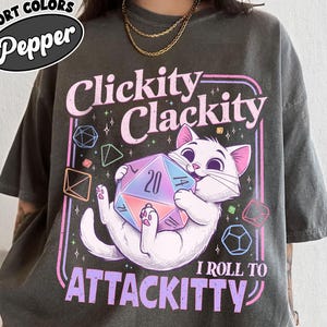 May include: Dark grey t-shirt with a graphic of a white cat holding a large, multi-sided die. Text reads "Clickity Clackity I Roll to Attackitty" in pink and purple. The shirt has a relaxed fit and the words "Comfort Colors Pepper" in a circle.