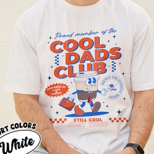 May include: White t-shirt with a retro graphic that reads "Proud member of the COOL DADS CLUB." The design features a coffee cup character carrying a briefcase. The text "STILL COOL JUST TIRED" is also included. The shirt is labeled "COMFORT COLORS White."