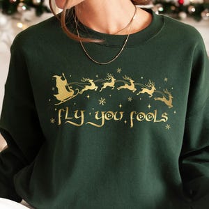 May include: A dark green sweatshirt with a gold graphic of Santa Claus in a sleigh pulled by reindeer, with the text "Fly you fools". The design includes snowflakes and stars, creating a festive holiday theme.