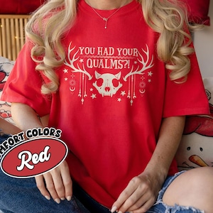 May include: A red short-sleeved t-shirt featuring a white graphic of a skull with antlers and the text "YOU HAD YOUR QUALMS?!". The design includes hanging ornaments, stars, and celestial symbols. A red oval label with the text "COMFORT COLORS Red" is visible.