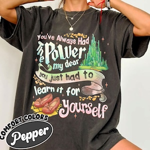 May include: Dark gray t-shirt with the text "You've Always Had Power my dear you just had to learn it for Yourself". The graphic includes a green castle, red shoes, and the words "Comfort Colors Pepper".