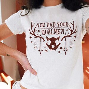 May include: A white short-sleeved t-shirt featuring a dark red graphic print. The design includes a central animal skull, large antlers, celestial symbols like stars, moons, and suns, and the text "YOU HAD YOUR QUALMS?!" in a bold, stylized font.