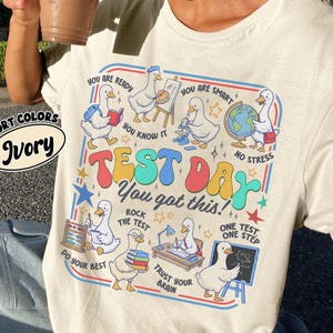 May include: Ivory-colored t-shirt with a colorful graphic that says "TEST DAY! You got this!" and features cartoon ducks. The shirt also has the words "YOU ARE READY", "YOU ARE SMART", "NO STRESS", "DO YOUR BEST", "ROCK THE TEST", "TRUST YOUR BRAIN", and "ONE TEST ONE STEP".