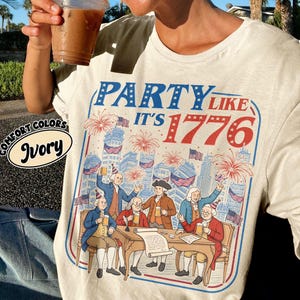 Puede incluir: Camiseta de color marfil con un gr&aacute;fico que dice "PARTY LIKE IT'S 1776" en azul y rojo. El gr&aacute;fico representa a un grupo de hombres con atuendo colonial celebrando con cerveza y fuegos artificiales. La camiseta tiene una etiqueta Comfort Colors.