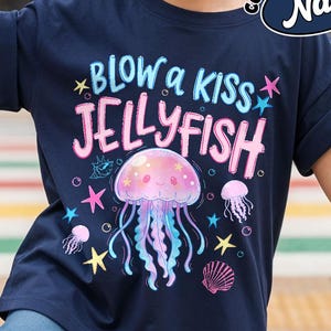 May include: Navy blue t-shirt with the text "BLOW a KISS JELLYFISH" and a colorful jellyfish graphic. The design includes pink, blue, and yellow accents, with additional starfish and seashell illustrations. The shirt has rolled-up sleeves.
