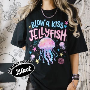 May include: Black t-shirt with a colorful jellyfish graphic and the text "BLOW a KISS JELLYFISH". The design includes stars, a seashell, and smaller jellyfish. The shirt is labeled "COMFORT COLORS Black".
