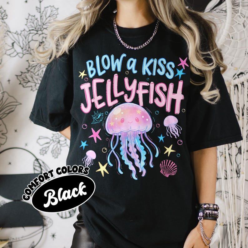 May include: Black t-shirt with a colorful jellyfish graphic and the text "BLOW a KISS JELLYFISH". The design includes stars, a seashell, and smaller jellyfish. The shirt is labeled "COMFORT COLORS Black".