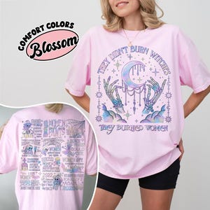 May include: A light pink t-shirt with the text "THEY DIDN'T BURN WITCHES THEY BURNED WOMEN" and a graphic of a crescent moon, hands, and stars. The shirt also has the text "COMFORT COLORS Blossom" on it.