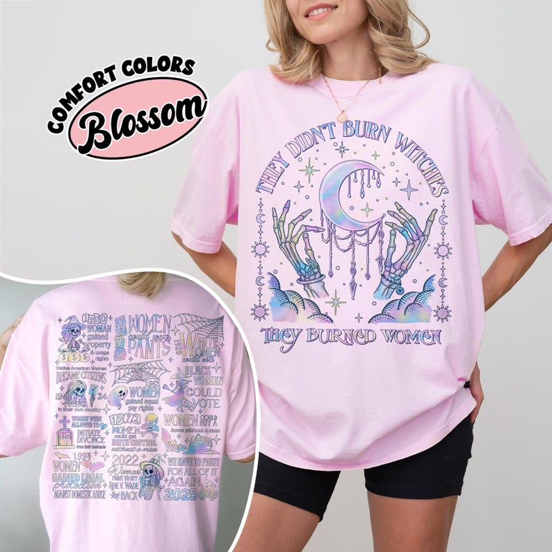 May include: A light pink t-shirt with the text "THEY DIDN'T BURN WITCHES THEY BURNED WOMEN" and a graphic of a crescent moon, hands, and stars. The shirt also has the text "COMFORT COLORS Blossom" on it.
