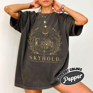 May include: A dark gray oversized t-shirt with a gold graphic design of a castle, moon, stars, and leaves. The text "SKYHOLD" and "THE PLACE WHERE THE SKY IS KEPT" is printed below the graphic. The shirt has a distressed look and is made by Comfort Colors Pepper.