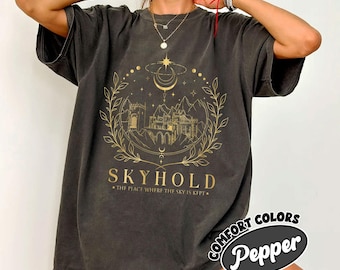 May include: A dark gray oversized t-shirt with a gold graphic design of a castle, moon, stars, and leaves. The text "SKYHOLD" and "THE PLACE WHERE THE SKY IS KEPT" is printed below the graphic. The shirt has a distressed look and is made by Comfort Colors Pepper.