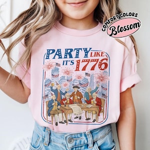 Puede incluir: Una camiseta rosa claro con un dise&ntilde;o gr&aacute;fico con el texto "PARTY LIKE IT'S 1776" en rojo y azul. El dise&ntilde;o muestra a hombres con ropa colonial celebrando con fuegos artificiales y banderas. La camiseta es de Comfort Colors.