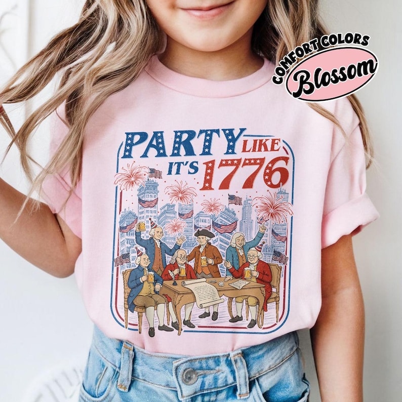 Puede incluir: Una camiseta rosa claro con un dise&ntilde;o gr&aacute;fico con el texto "PARTY LIKE IT'S 1776" en rojo y azul. El dise&ntilde;o muestra a hombres con ropa colonial celebrando con fuegos artificiales y banderas. La camiseta es de Comfort Colors.