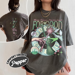 May include: Dark grey t-shirt with a whimsical design featuring a clock, hands, a witch's hat, and the text "FOR GOOD." The back of the shirt has a matching design. The shirt is labeled "Comfort Colors Pepper."