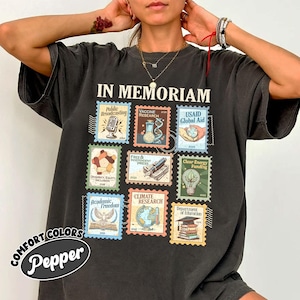 May include: Dark gray t-shirt with the words "IN MEMORIAM" above a collection of colorful postage stamp designs. The stamps feature various themes, including public broadcasting, vaccine research, and climate research. The shirt also has the words "COMFORT COLORS" and "Pepper" printed on it.