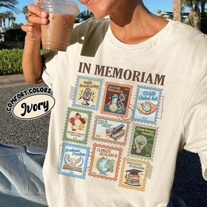 May include: Ivory-colored t-shirt with the words "IN MEMORIAM" above a collection of postage stamp-style graphics. The stamps depict themes like "Public Broadcasting," "Vaccine Research," and "Climate Research." The shirt also features the words "COMFORT COLORS" and "Ivory."