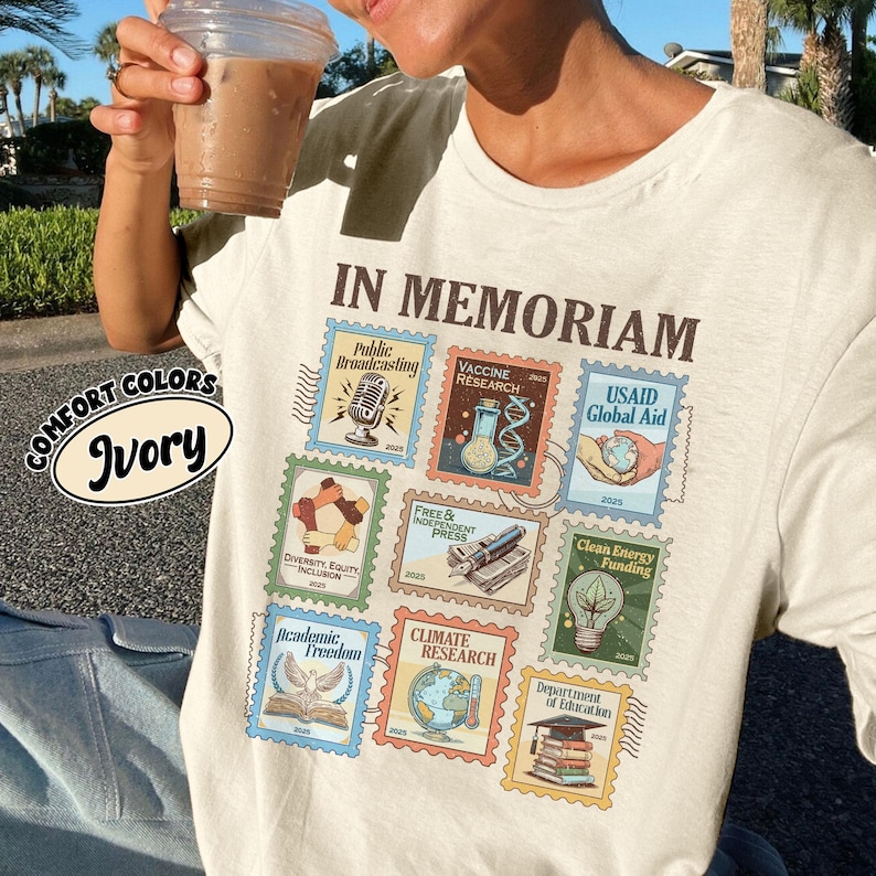 May include: Ivory-colored t-shirt with the words "IN MEMORIAM" above a collection of postage stamp-style graphics. The stamps depict themes like "Public Broadcasting," "Vaccine Research," and "Climate Research." The shirt also features the words "COMFORT COLORS" and "Ivory."